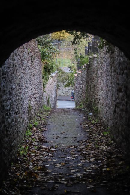 A narrow, paved pathway viewed from beneath a dark stone archway, leading out to a street beyond. The sides of the pathway are flanked by tall, weathered brick and stone walls covered with moss and cl