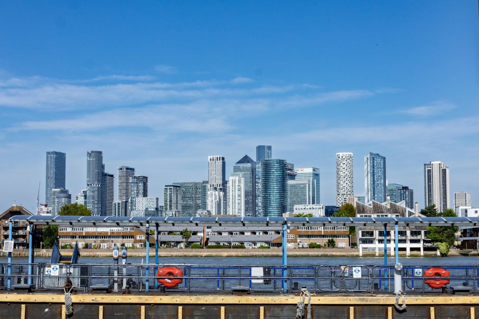 A wide view of a modern urban skyline featuring numerous high-rise office buildings and residential towers with reflective glass facades, set against a bright blue sky with scattered clouds. In the fo