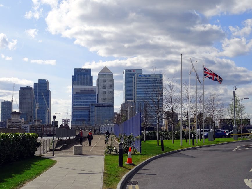 A wide view of a modern urban skyline featuring numerous high-rise office buildings and residential towers with reflective glass facades, set against a bright blue sky with scattered clouds. In the fo
