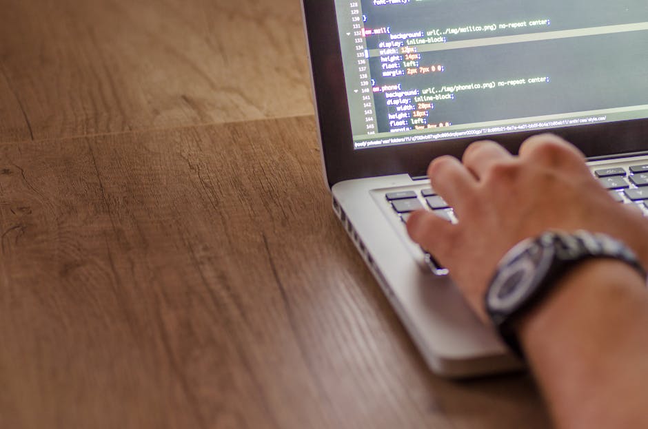 A close-up of a person's hand with a black and silver watch, typing on a laptop placed on a wooden desk. The laptop screen displays lines of code with various colors, indicating programming or web dev